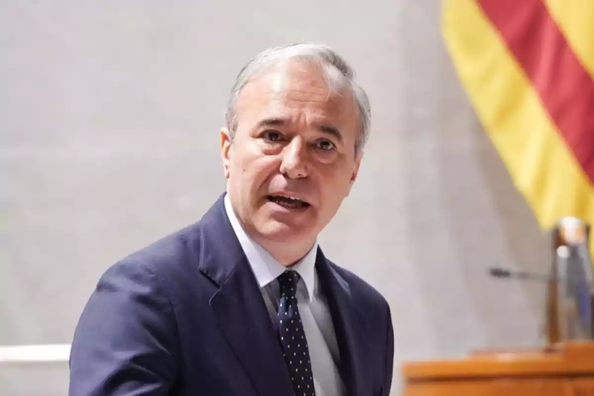 Hombre de cabello canoso y traje oscuro hablando frente a un micrófono con una bandera en el fondo.