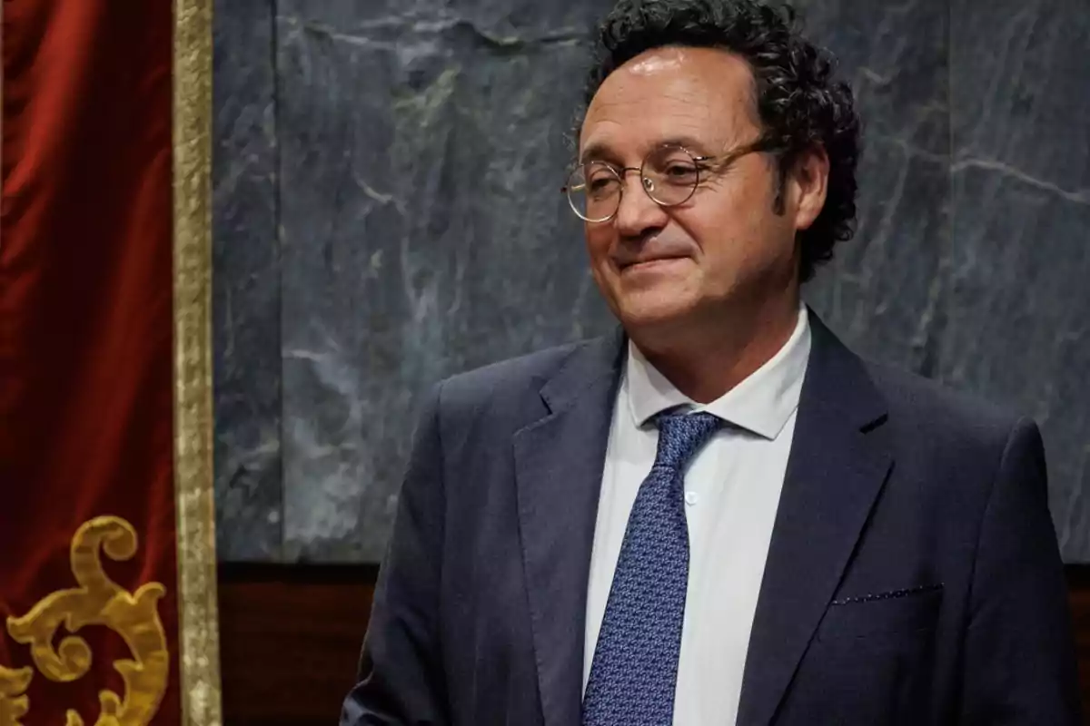 Hombre con gafas y traje azul sonriendo frente a un fondo de mármol y una cortina roja decorativa.