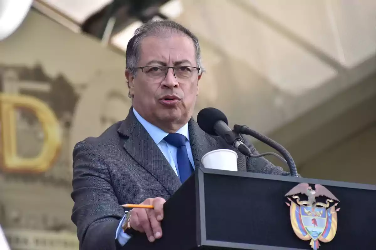 Un hombre con gafas y traje gris habla en un podio con un escudo, sosteniendo un lápiz y un vaso de papel.