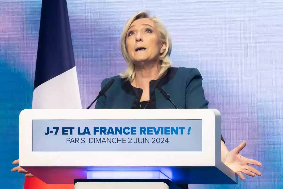 Una mujer rubia está dando un discurso en un podio con un micrófono, con una bandera de Francia a su lado y un cartel que dice "J-7 ET LA FRANCE REVIENT! PARIS, DIMANCHE 2 JUIN 2024".
