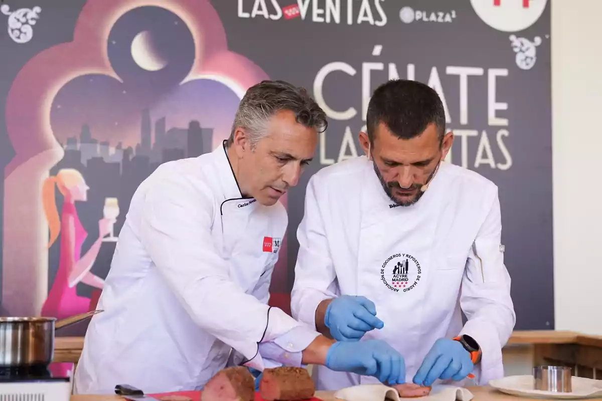 Dos chefs con chaquetas blancas y guantes azules preparan un plato en un evento culinario, con un fondo que muestra un cartel de "Cénate Las Ventas".