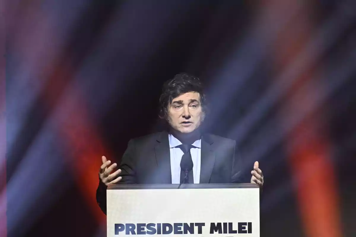 Hombre de traje oscuro hablando en un podio con el texto "PRESIDENT MILEI" iluminado por luces de colores.