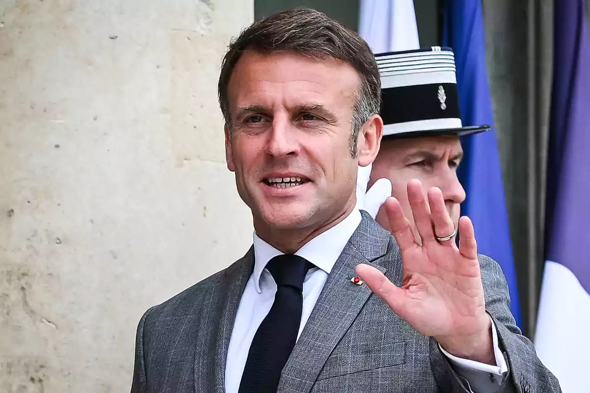 Hombre con traje gris y corbata negra saludando con la mano levantada, con un oficial de fondo.
