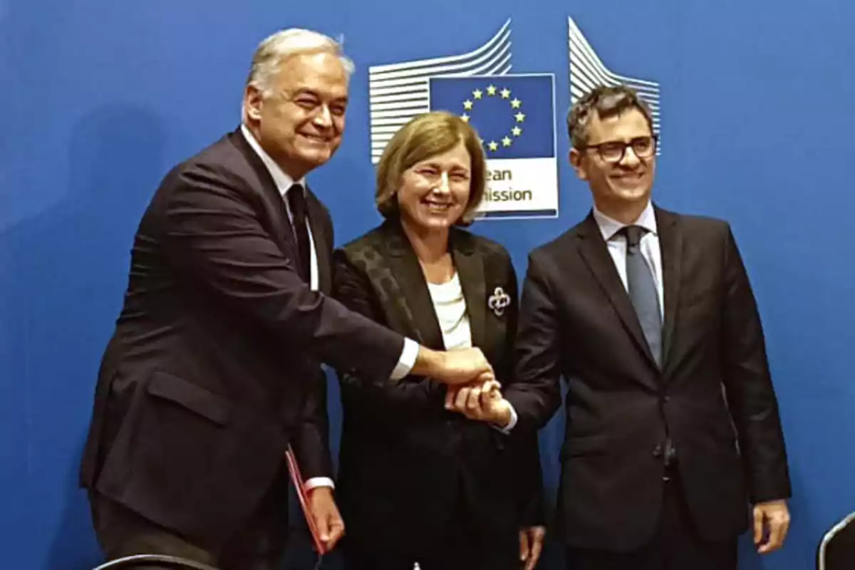 Tres personas en trajes formales posan frente a un fondo azul con el logotipo de la Comisión Europea.