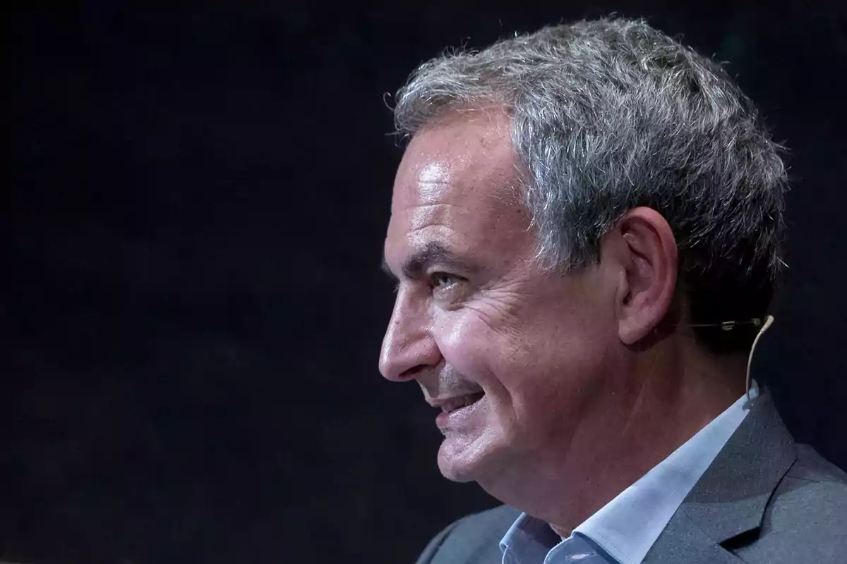 Man in profile with gray hair and microphone in his ear, dark background.