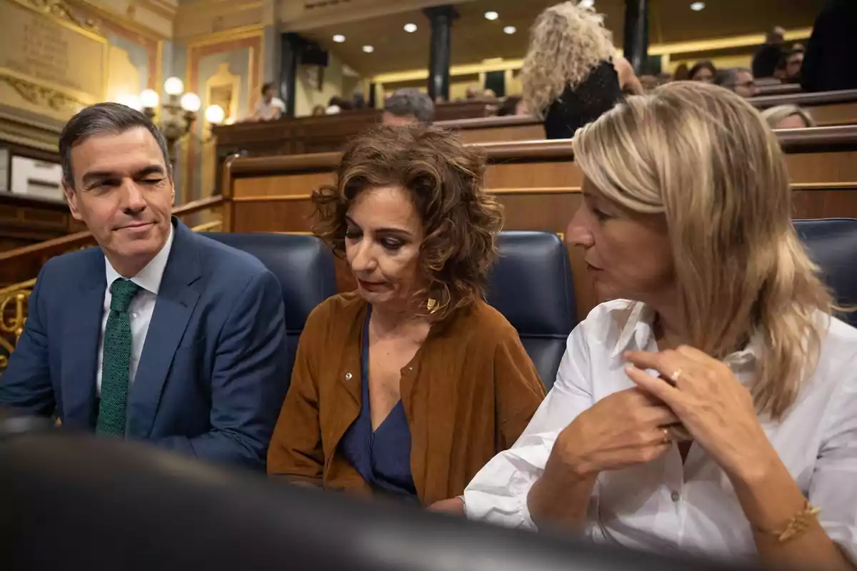 Tres personas sentadas en un entorno formal conversan entre ellas mientras una de ellas parece estar tomando notas o revisando algo.