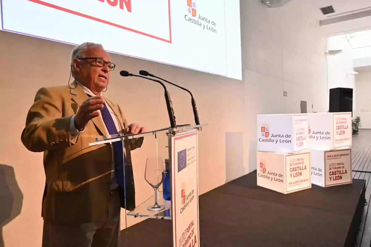 Un hombre mayor con gafas y traje marrón claro está hablando en un podio con micrófonos, en un evento de la Junta de Castilla y León.