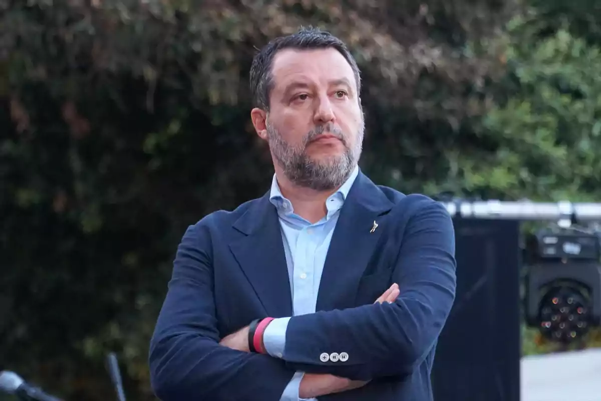 Un hombre con barba y cabello oscuro viste un traje azul y camisa clara mientras cruza los brazos en un entorno al aire libre.