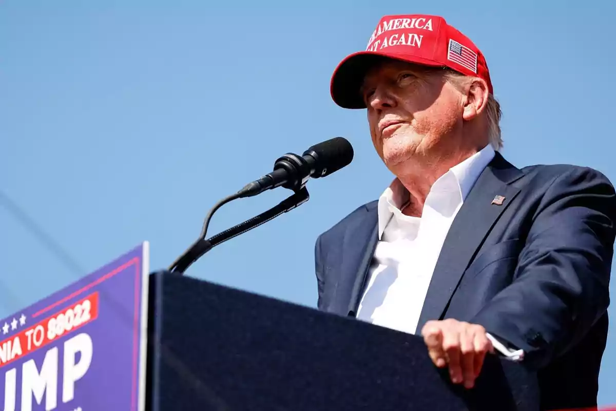 Un hombre con una gorra roja que dice "Make America Great Again" habla en un mitin frente a un micrófono.