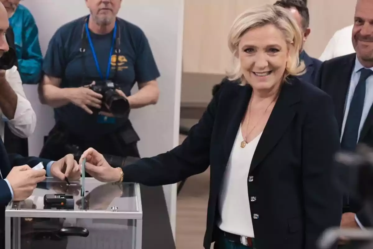 Una mujer rubia con chaqueta oscura y camisa blanca está sonriendo mientras deposita un voto en una urna transparente, rodeada de personas, incluyendo un fotógrafo con una cámara.