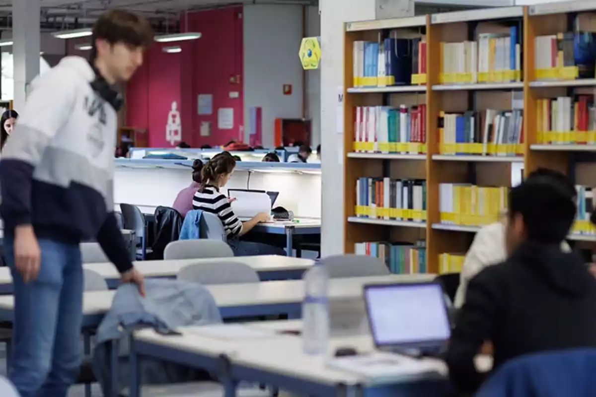 Estudiantes trabajando y estudiando en una biblioteca con estanterías llenas de libros y mesas con computadoras portátiles.