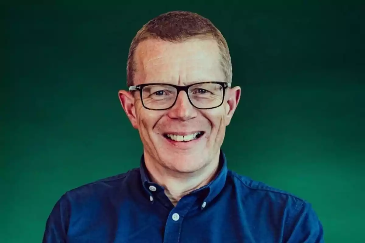 Hombre con gafas y camisa azul sonriendo frente a un fondo verde.