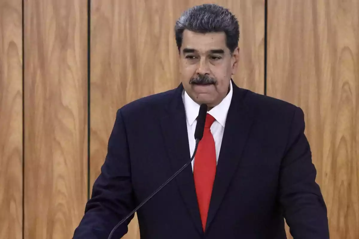 Hombre con traje oscuro y corbata roja hablando frente a un micrófono con fondo de madera.