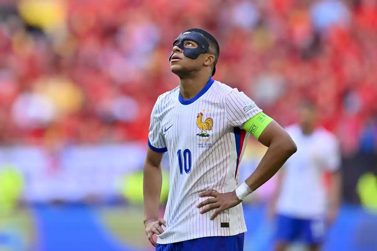 Jugador de fútbol con máscara negra y camiseta blanca con rayas rojas y azules, mirando hacia arriba en un estadio.