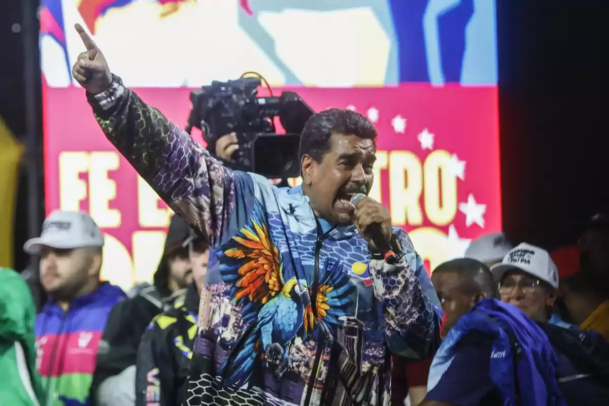 Un hombre con una chaqueta colorida y un micrófono en la mano, levantando el brazo mientras habla frente a una multitud y una pantalla con texto en el fondo.