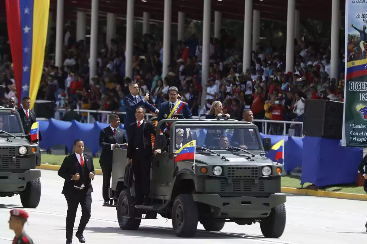 Desfile militar en Venezuela con vehículos militares y personas en uniforme, con una multitud observando desde las gradas y banderas venezolanas decorando el lugar.