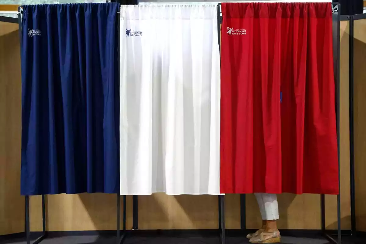 Cabinas de votación con cortinas de colores azul, blanco y rojo, con una persona parcialmente visible detrás de la cortina roja.