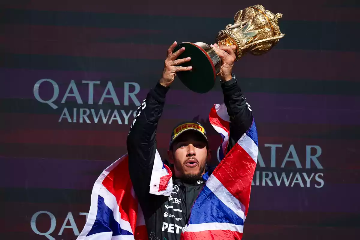 Piloto de carreras sosteniendo un trofeo dorado con una bandera británica sobre sus hombros y un fondo de Qatar Airways.