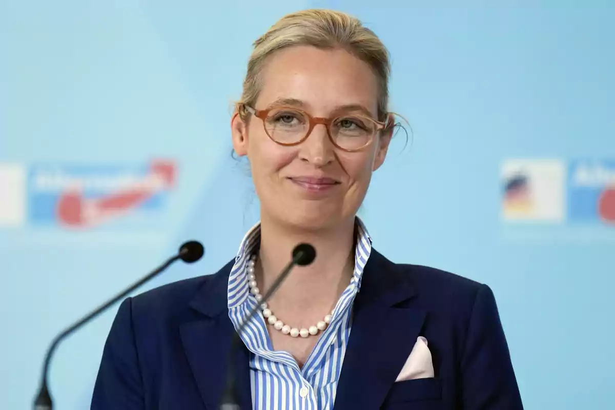 A person with glasses and a blue suit speaks in front of a microphone with a light blue background.