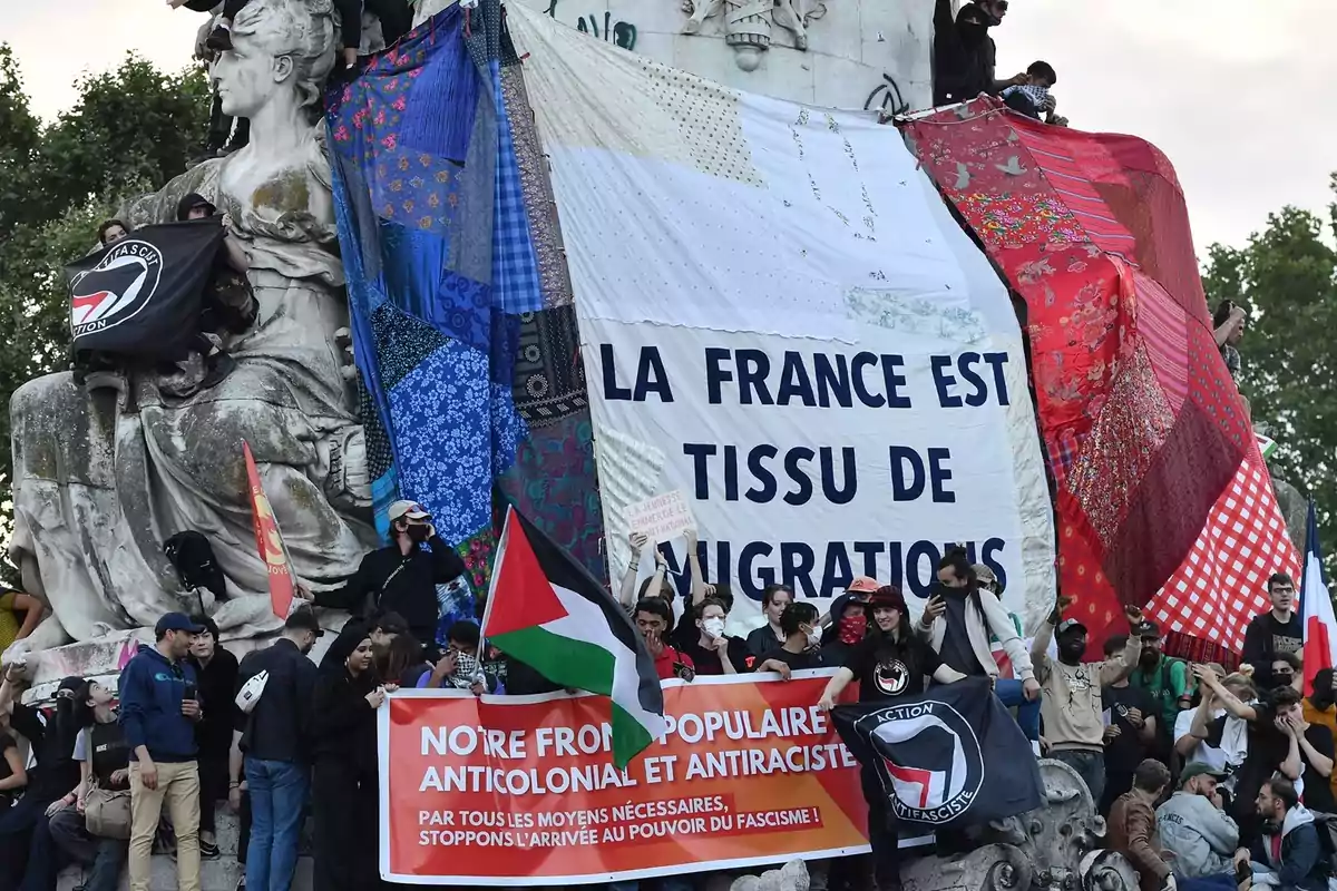 Manifestantes se reúnen alrededor de una estatua, con pancartas y banderas, destacando mensajes en contra del fascismo y en apoyo a la diversidad y la inmigración en Francia.