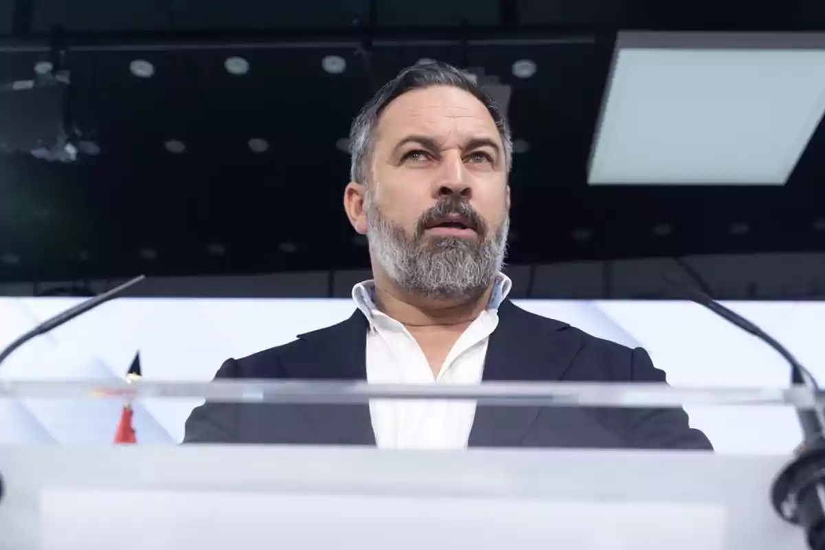 Hombre de barba y cabello corto, vestido con camisa blanca y chaqueta oscura, hablando en un podio con micrófonos frente a un fondo oscuro con luces.