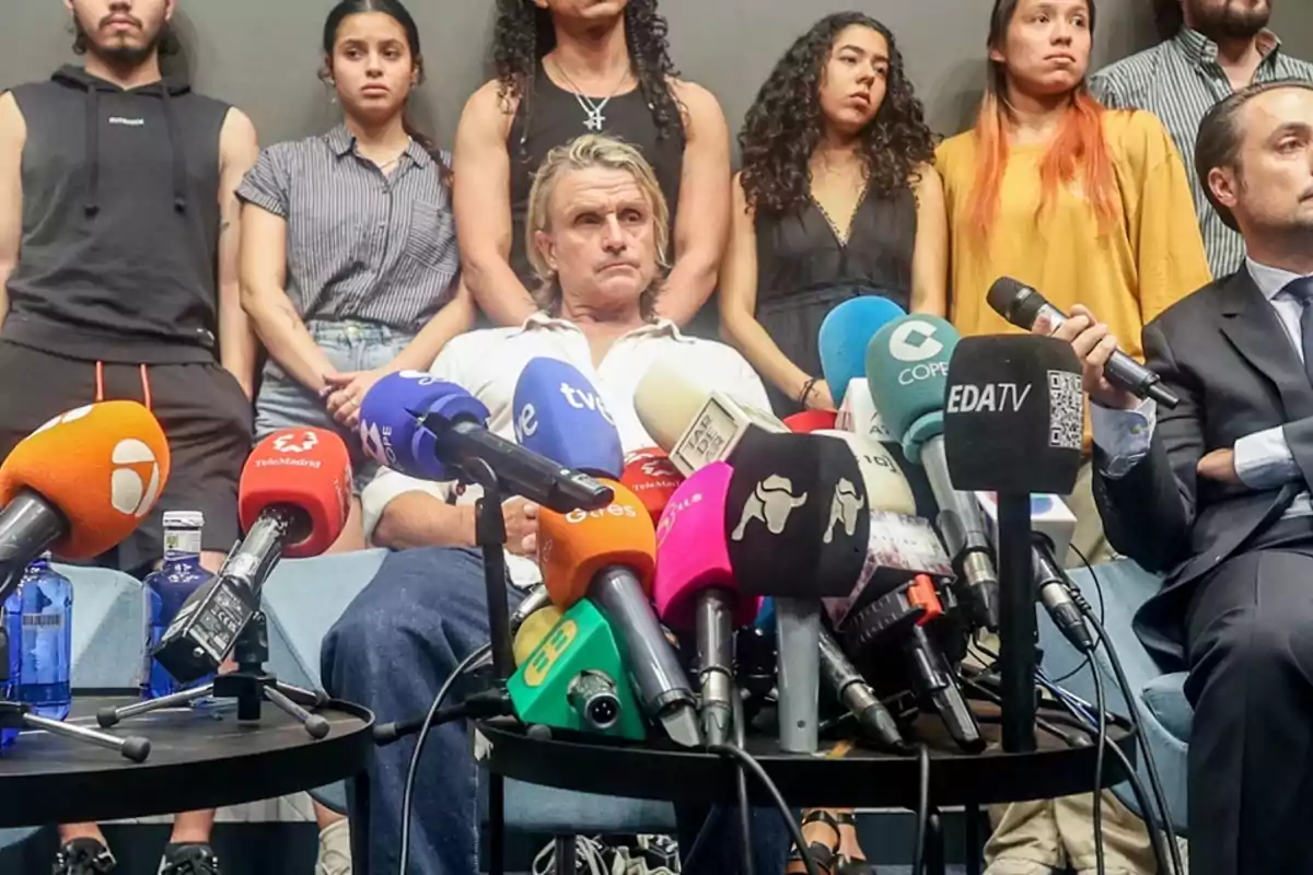 Personas sentadas y de pie en una conferencia de prensa con múltiples micrófonos de diferentes medios de comunicación en primer plano.