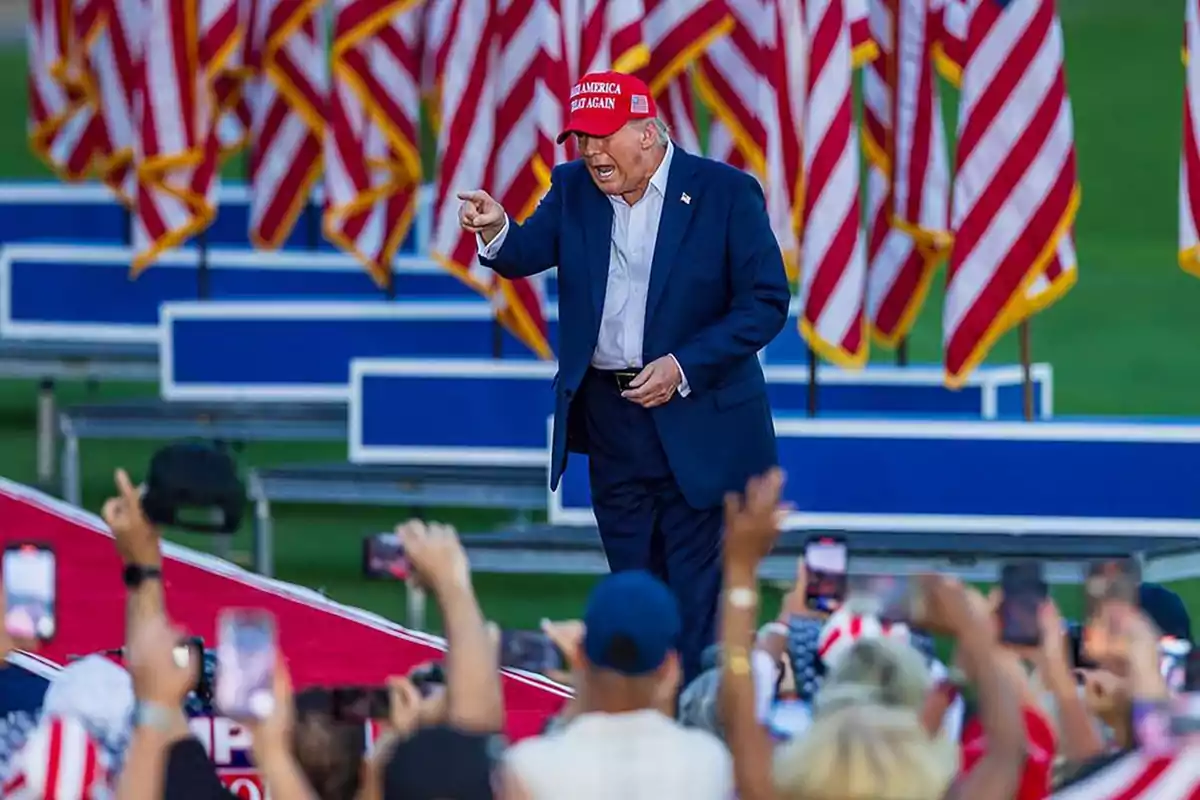 Persona con gorra roja y traje azul en un evento al aire libre con banderas estadounidenses de fondo y una multitud tomando fotos.