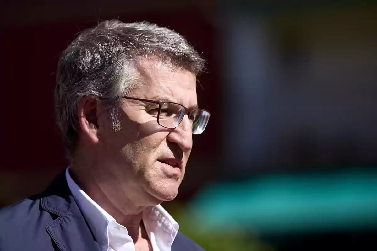 Hombre con gafas y cabello canoso, vestido con una chaqueta azul y camisa blanca, mirando hacia la derecha con expresión seria.