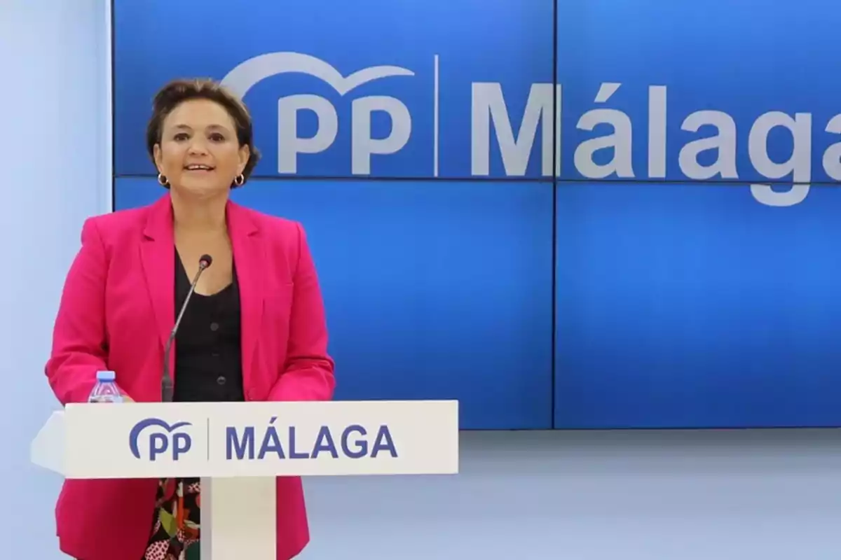 Una persona con chaqueta rosa habla en un podio con el logotipo del PP y la palabra "Málaga" en el fondo azul.