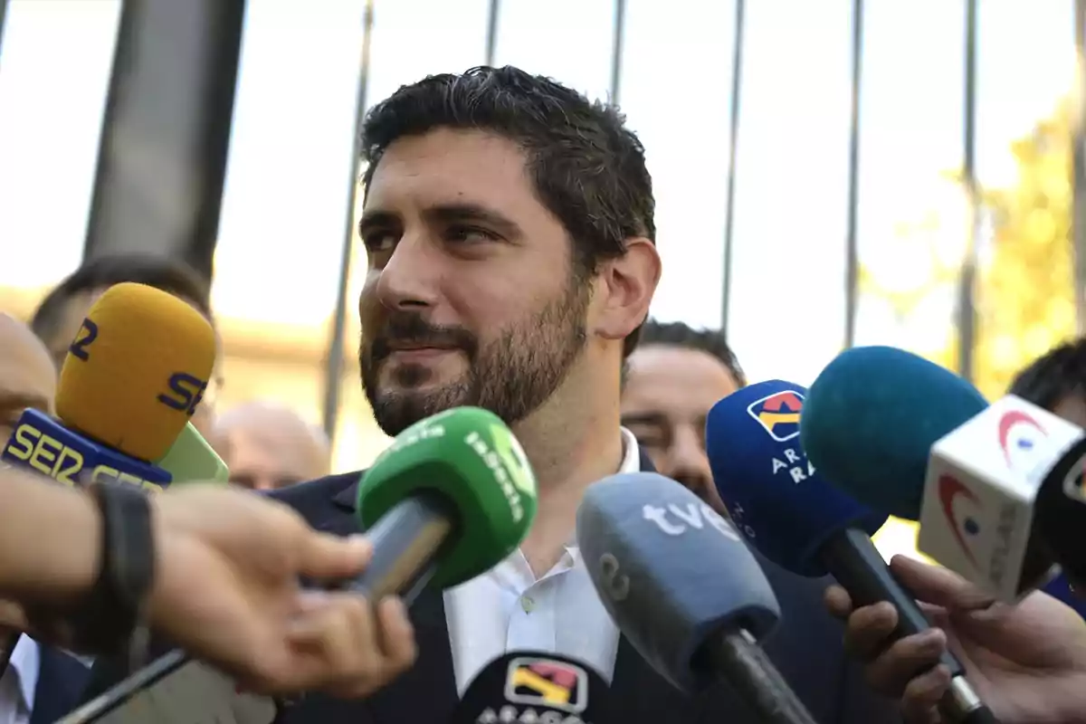 Un hombre con barba es entrevistado por varios medios de comunicación, rodeado de micrófonos de diferentes cadenas de televisión y radio.