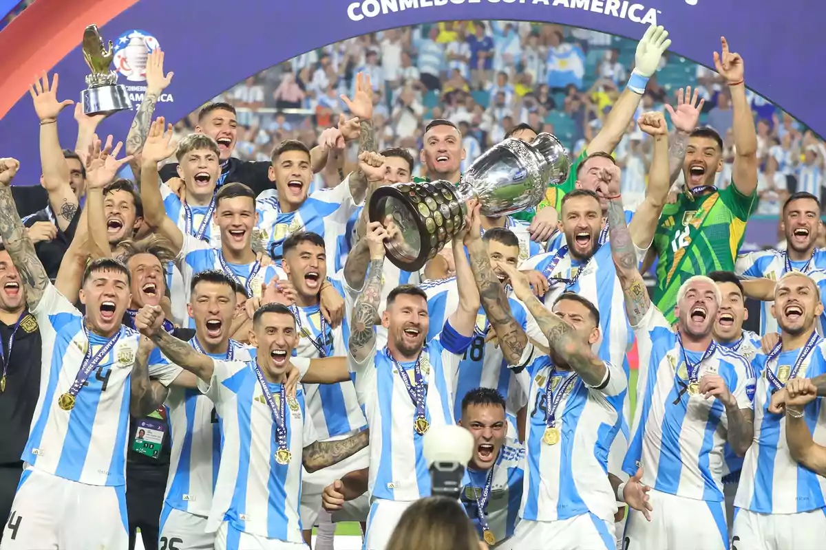 Jugadores de un equipo de fútbol celebran con entusiasmo mientras levantan un trofeo, vistiendo uniformes a rayas celestes y blancas, con medallas colgando de sus cuellos y rodeados de un ambiente festivo.