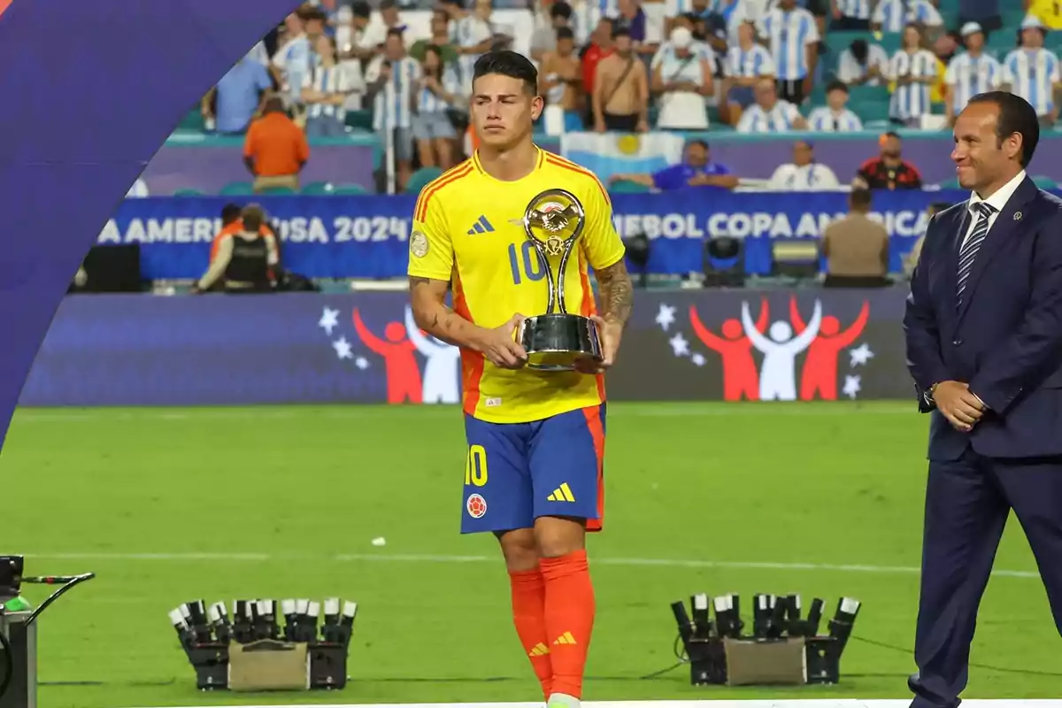 Jugador de fútbol con uniforme amarillo y azul sosteniendo un trofeo en un estadio, con un hombre de traje a su lado y público en el fondo.