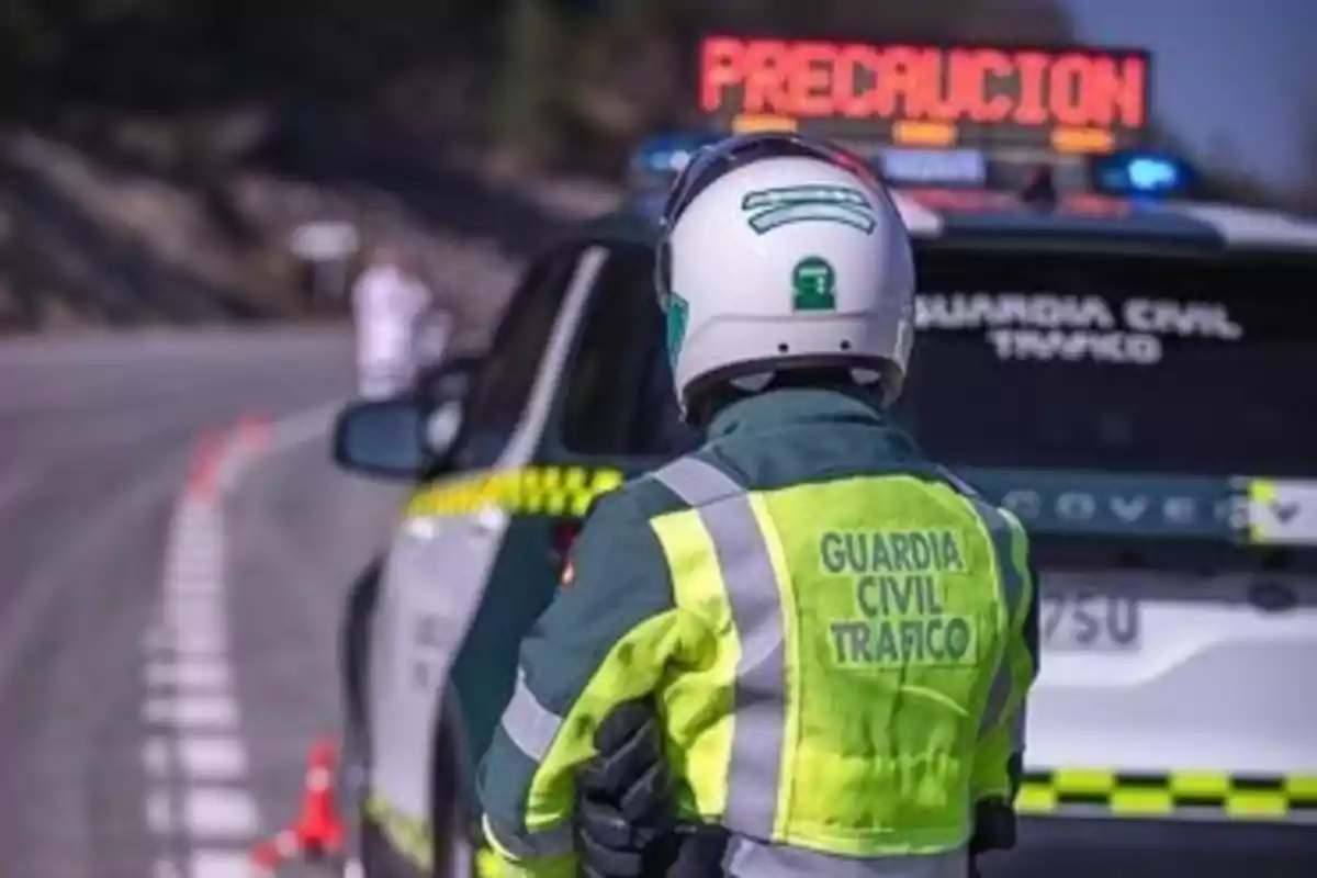 Un agente de la Guardia Civil de Tráfico con chaleco reflectante y casco observa una carretera mientras un coche patrulla está estacionado con un letrero de precaución iluminado. Un agente de la Guardia Civil de Tráfico con chaleco reflectante y casco observa una carretera mientras un coche patrulla está estacionado con un letrero de precaución iluminado.
