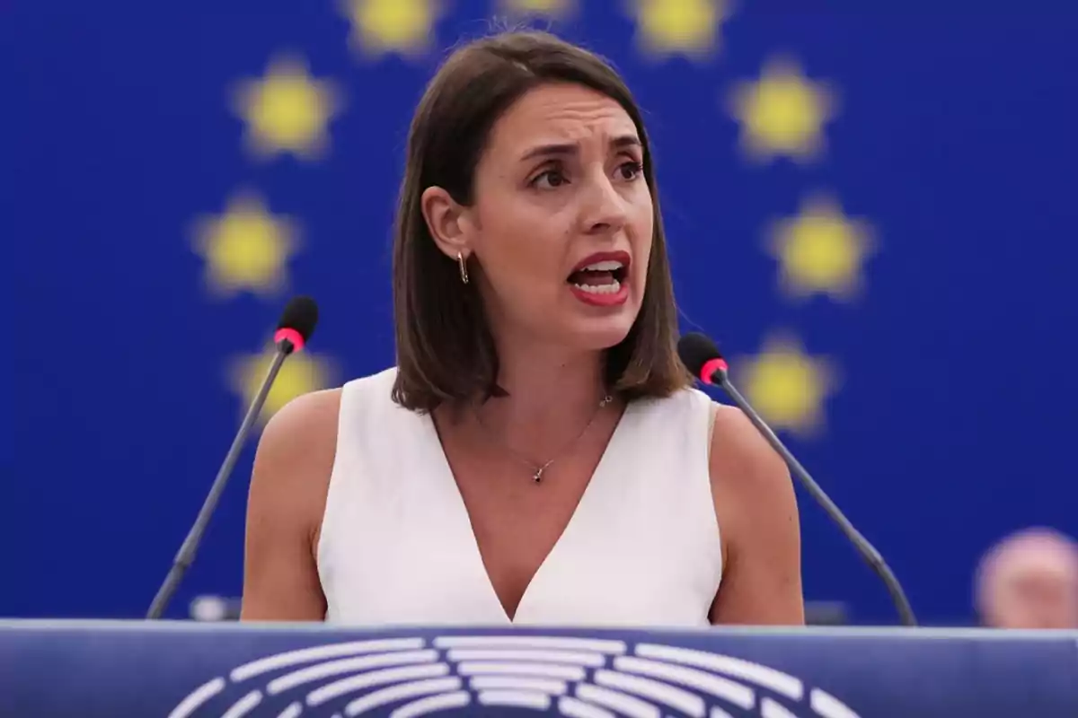 Mujer hablando en un podio con el fondo de la bandera de la Unión Europea.