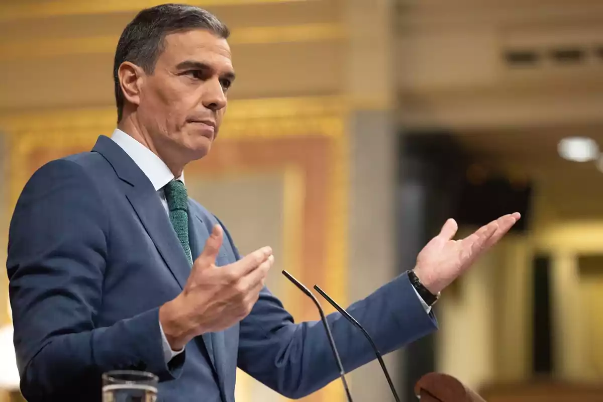 Hombre de traje azul y corbata verde hablando en un podio con gestos expresivos.