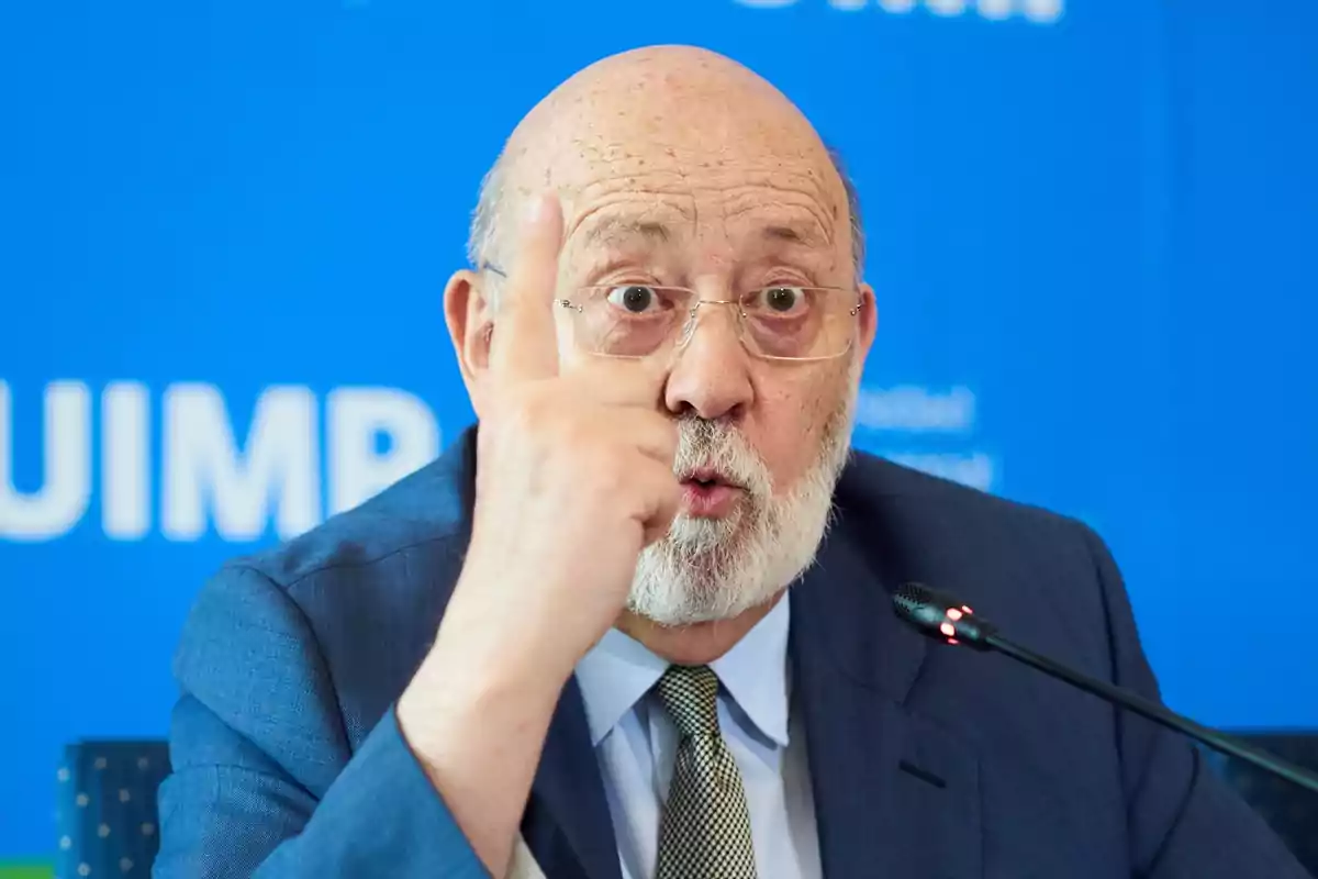 Hombre mayor con barba y gafas hablando en una conferencia y levantando el dedo índice mientras gesticula frente a un fondo azul Hombre mayor con barba y gafas hablando en una conferencia y levantando el dedo índice mientras gesticula frente a un fondo azul