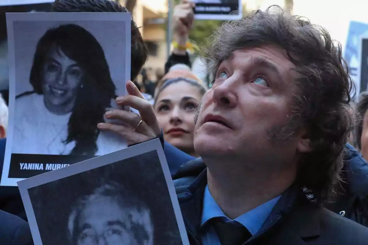 Una persona con cabello rizado y expresión seria sostiene una fotografía en blanco y negro de una mujer, mientras otras personas en el fondo también sostienen imágenes similares.