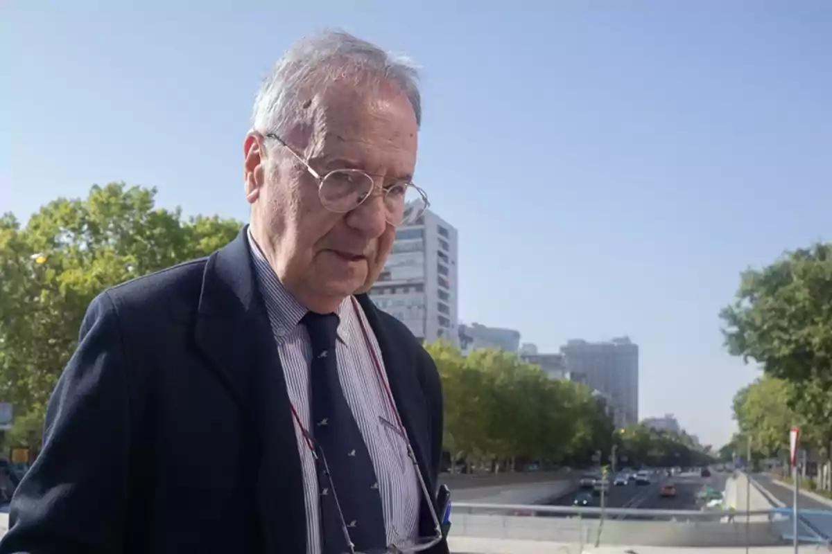 Un hombre mayor con gafas y traje camina por una calle urbana con árboles y edificios al fondo.