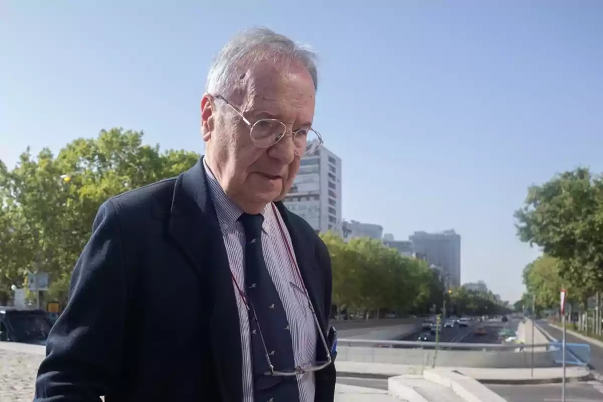 Hombre mayor con gafas y traje caminando al aire libre en una ciudad con edificios y árboles al fondo.