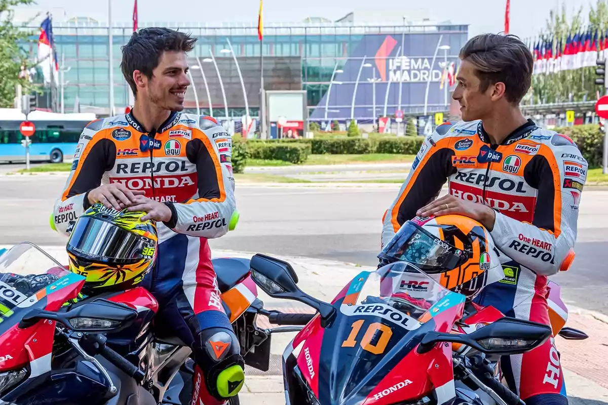 Dos motociclistas con trajes de carreras de Repsol Honda conversan mientras están sentados en sus motocicletas frente a un edificio con el letrero "IFEMA Madrid".