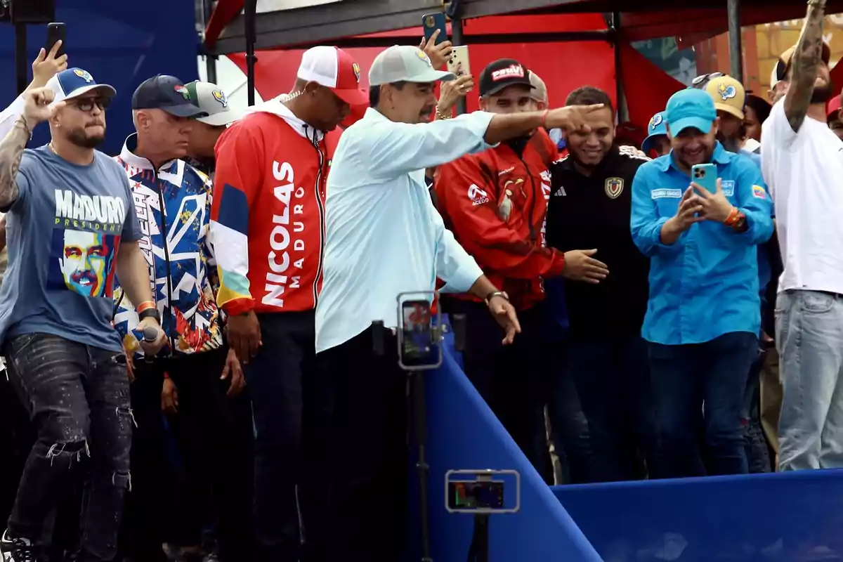 Un grupo de personas, algunas con ropa que tiene el nombre "Maduro", están reunidas en un evento al aire libre, algunas de ellas están señalando y otras tomando fotos con sus teléfonos.