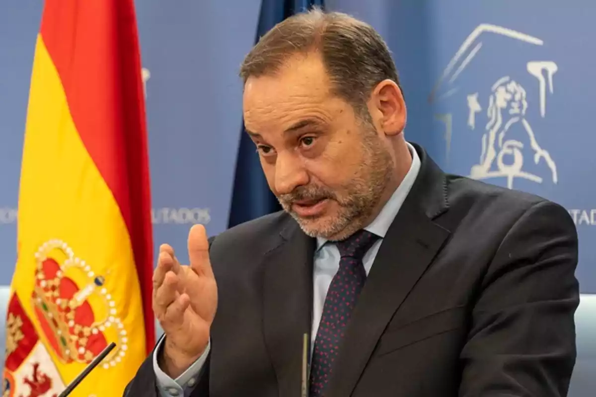 Un hombre en traje hablando en una conferencia de prensa con una bandera de España al fondo.