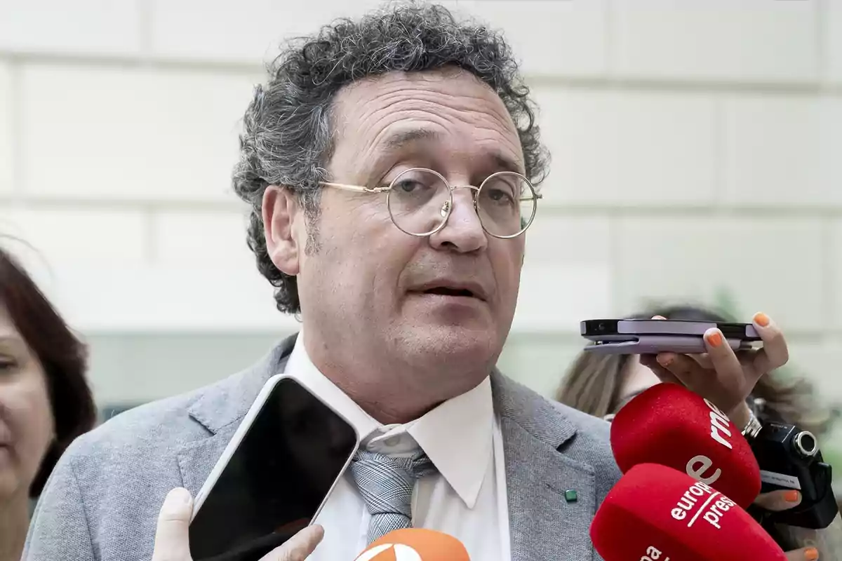 Hombre con gafas y cabello rizado hablando frente a varios micrófonos de prensa.