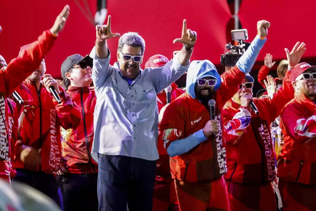 Un grupo de personas vestidas de rojo y usando gafas de sol, algunas con micrófonos, haciendo gestos con las manos en un evento con fondo rojo.