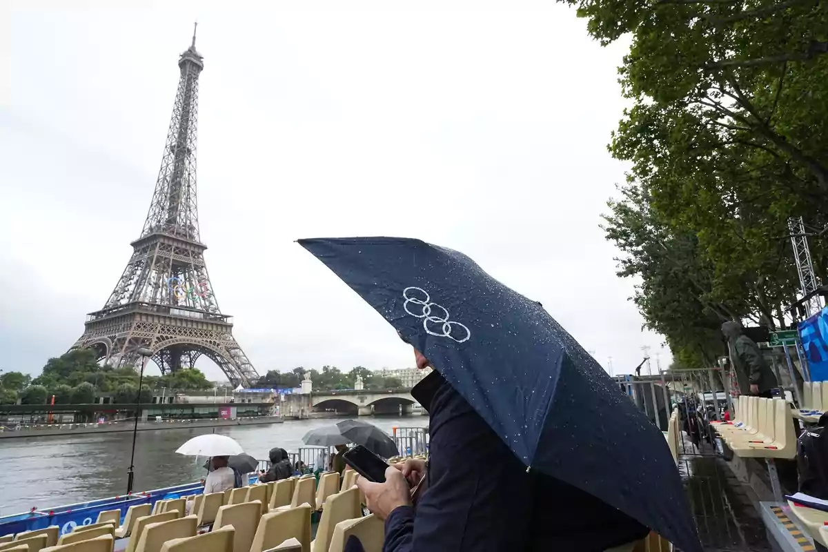 Persona con paraguas azul con el logo olímpico frente a la Torre Eiffel en un día lluvioso.