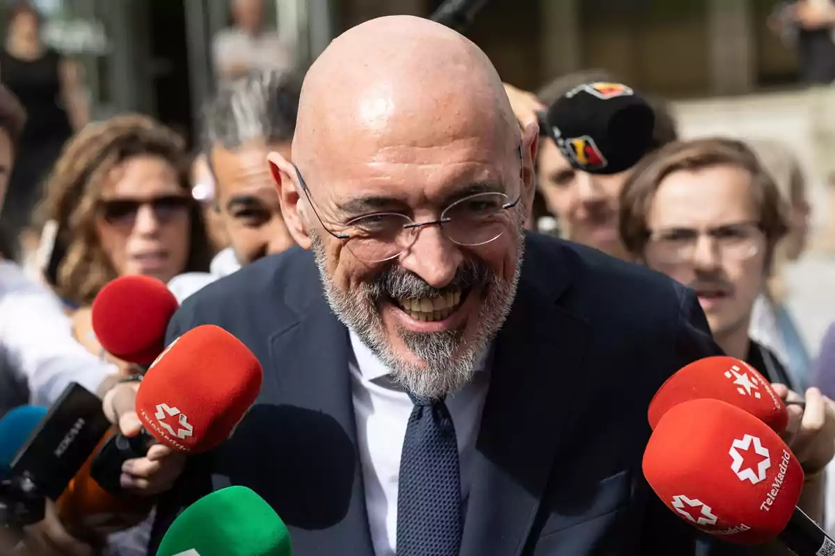 Un hombre calvo con barba y gafas sonríe mientras es rodeado por periodistas que sostienen micrófonos de diferentes colores.