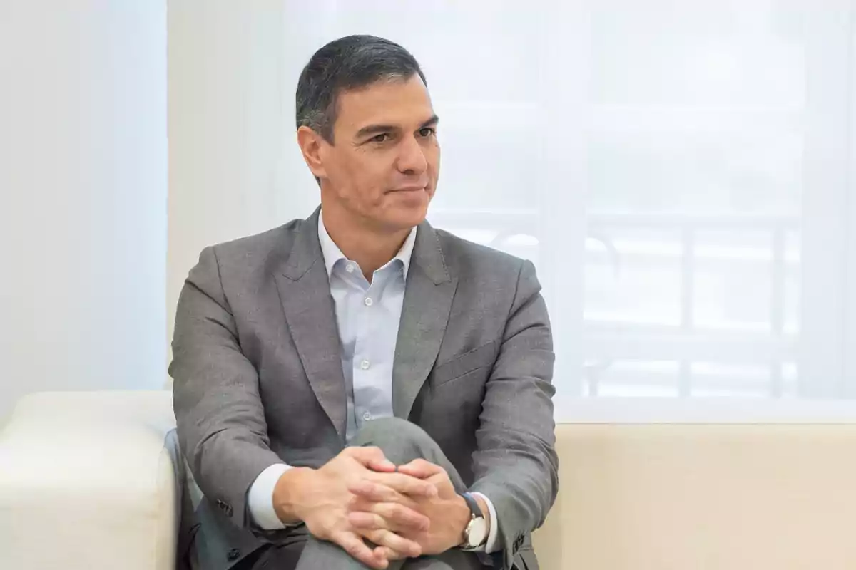 Un hombre con traje gris y camisa blanca está sentado en un sofá claro con las manos entrelazadas.