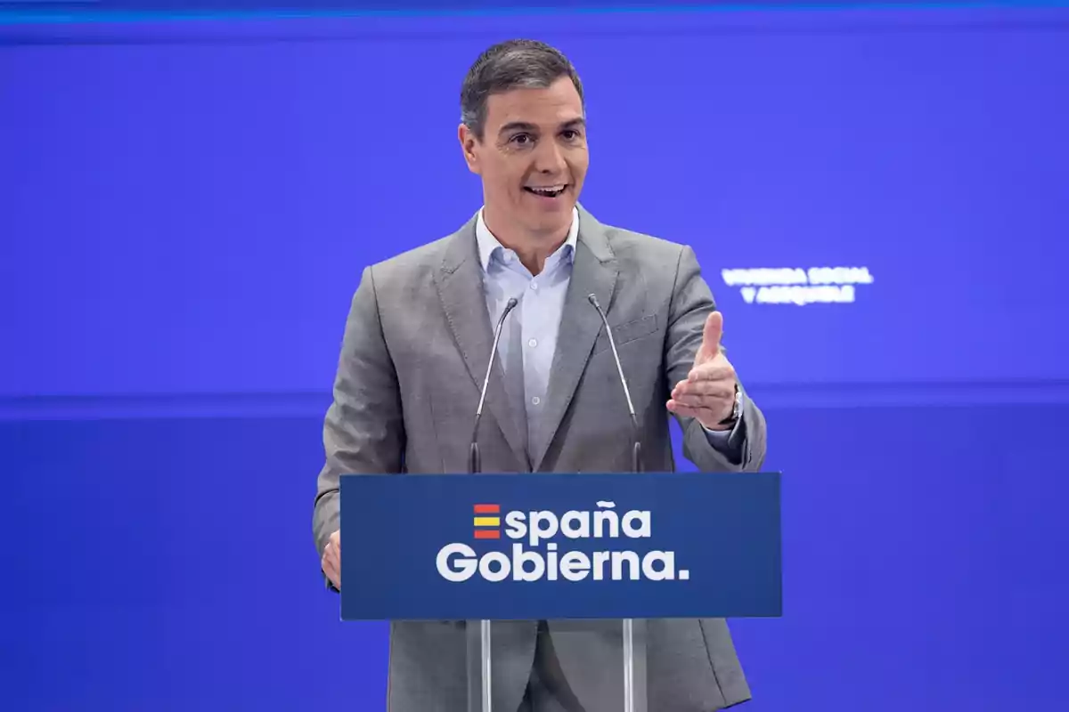 Un hombre en traje gris hablando en un podio azul con el texto "España Gobierna" en un fondo azul.