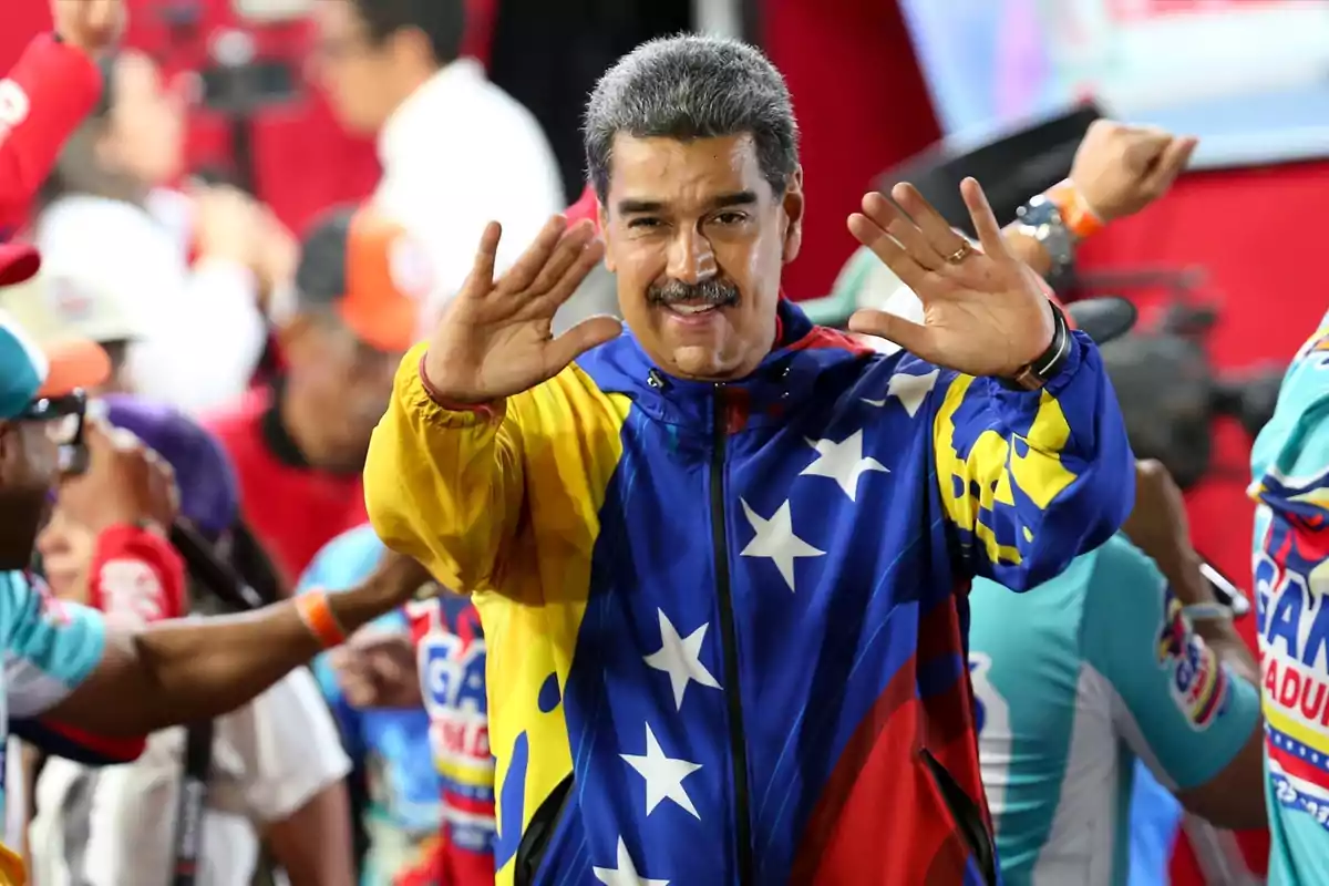 Un hombre con una chaqueta con los colores de la bandera de Venezuela saluda con ambas manos levantadas en un evento público.