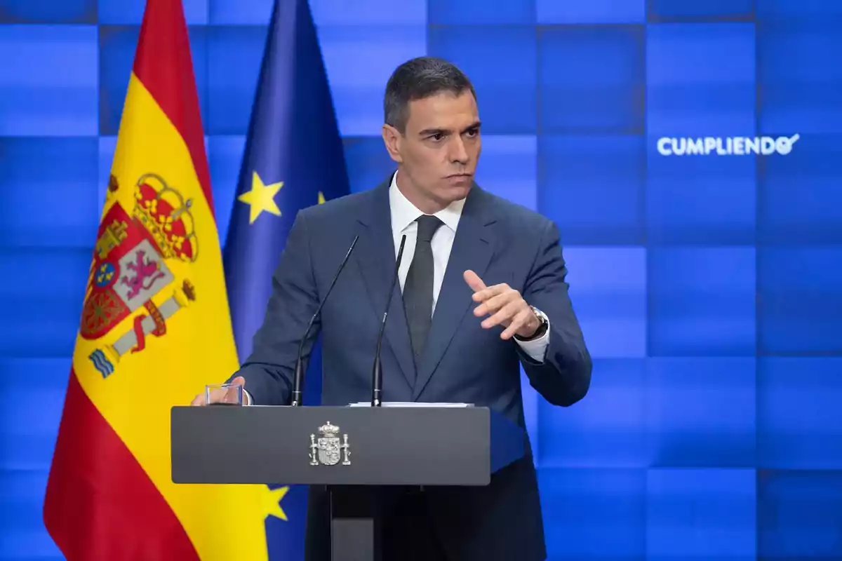 Un hombre en traje dando un discurso frente a un podio con las banderas de España y la Unión Europea de fondo.